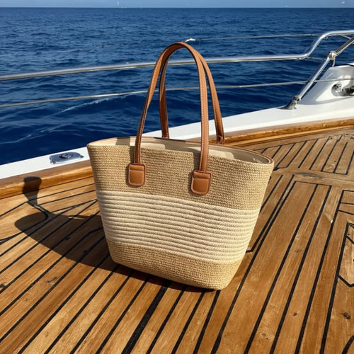 Bolsa feminina de palha bicolor com alças marrons sobre deck de barco, estilo náutico.